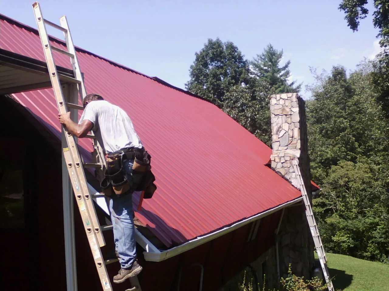 Roofer on a ladder beside a red metal roof and stone chimney — Soffit & Fascia Repair by Roofing Brothers in Mcleod